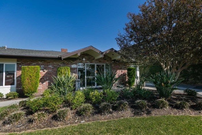 A mid-century style house with well-maintained landscaping, featuring bushes and trees under a clear blue sky.