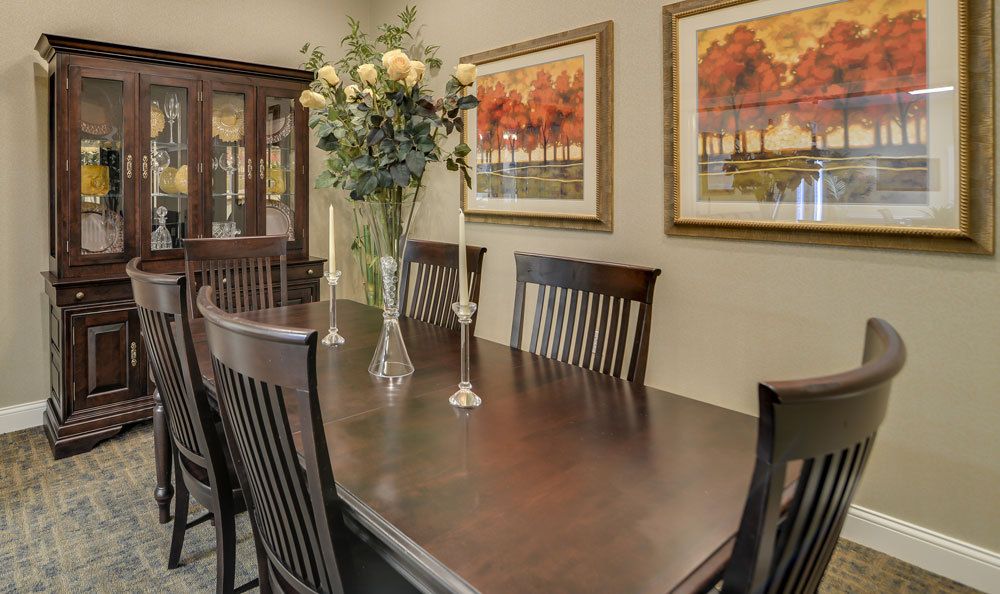 A well-decorated dining room featuring a dark wood table with matching chairs, adorned with a vase of flowers and candles. The walls are decorated with colorful landscape paintings.
