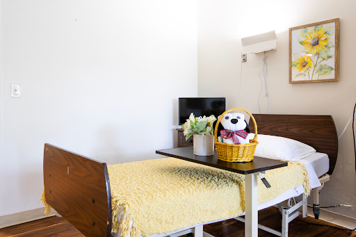 A cozy hospital room featuring a patient bed adorned with a yellow blanket, a small table with a decorative basket and stuffed animal, and a TV in the background.