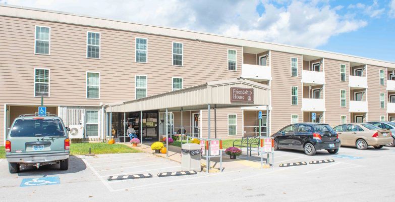 The image shows the exterior entrance of a hotel with a parking lot in front, taken during the day.