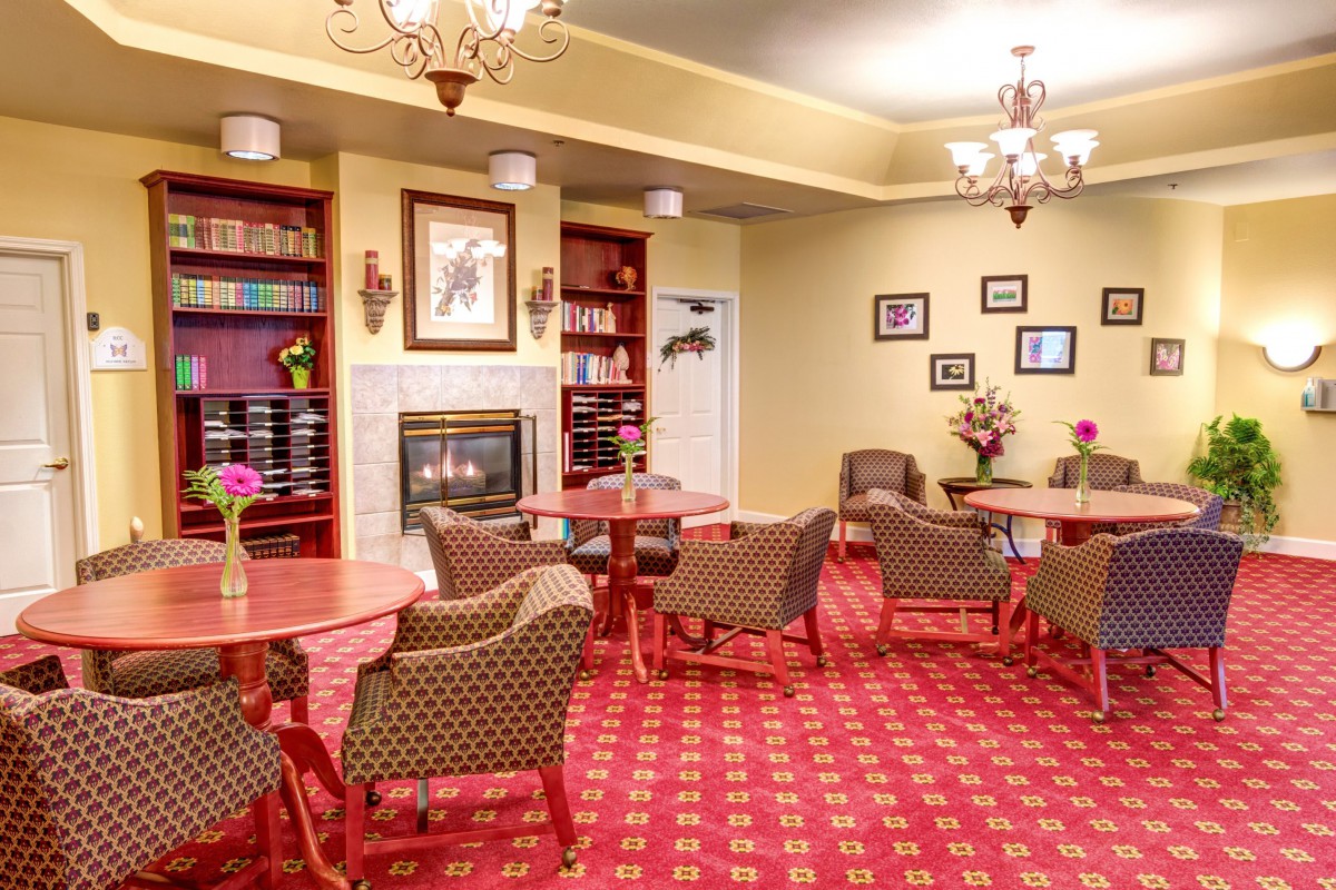 A cozy interior space with red carpet, featuring a fireplace, tables and chairs, bookshelves, and chandeliers.