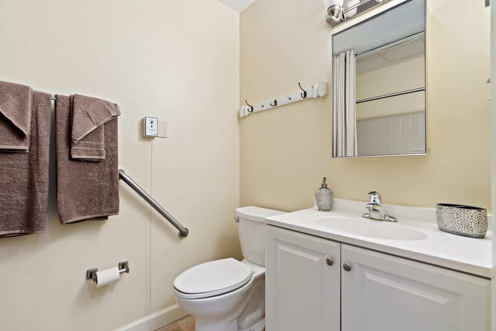 A small, clean bathroom with a toilet, sink, and mirror, featuring a towel rack and grab bar for accessibility.