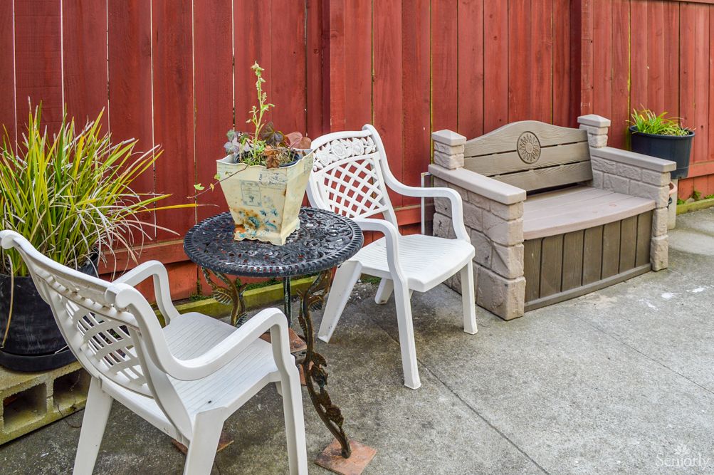 An outdoor patio setup with white plastic chairs, a table holding a planter, and a bench against a red wooden fence.