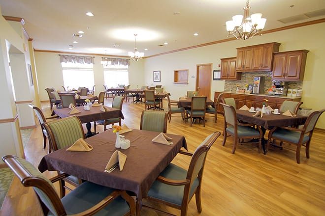 A spacious dining room with neatly arranged tables and chairs, featuring elegant chandeliers and a warm wooden floor.
