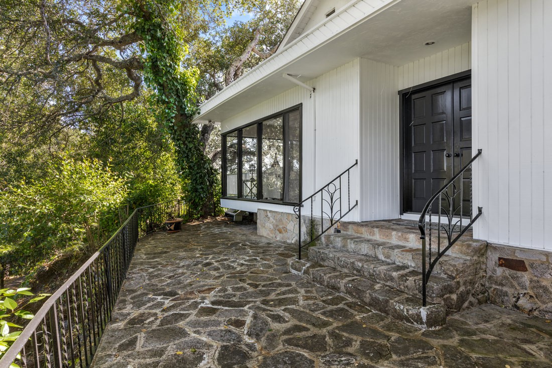 A view of a house with a stone patio and large windows surrounded by trees.