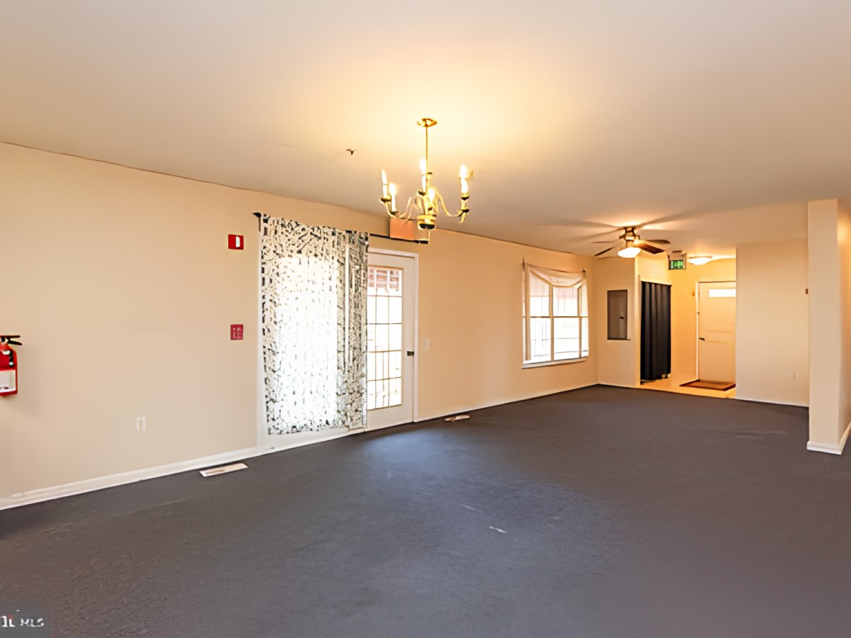 An empty room with carpet flooring, featuring a chandelier and ceiling fan, with windows and a doorway.