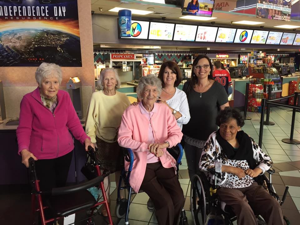 A group of people, including seniors in wheelchairs, smiling inside a cinema.