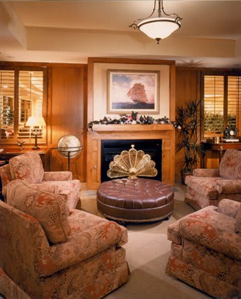 A cozy living room with plush armchairs, a central coffee table, and a decorative fireplace adorned with artwork and warm wood paneling.