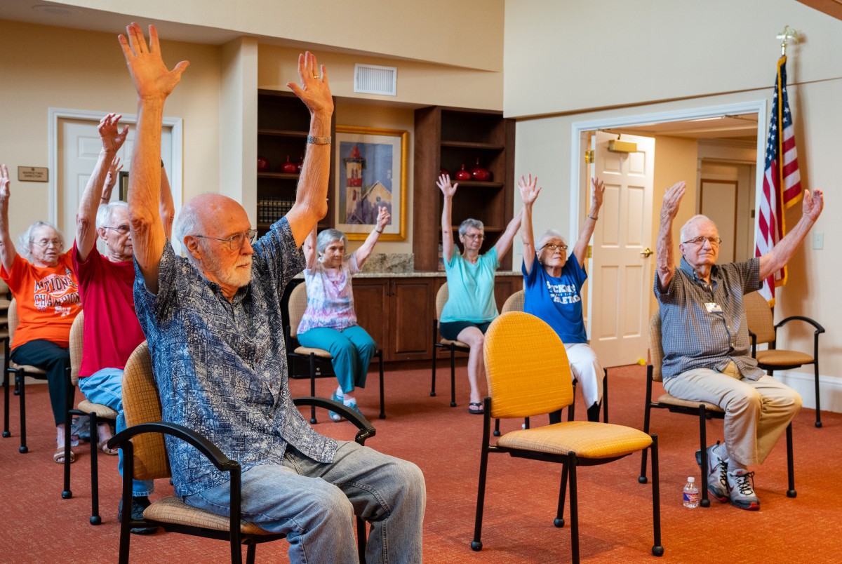 A group of seniors participating in a seated exercise session, raising their arms together in an indoor setting.