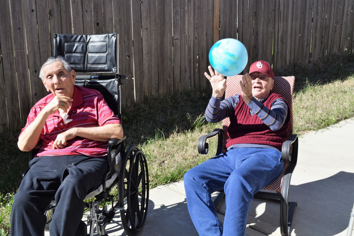 Two elderly individuals enjoying a sunny day outdoors, one in a wheelchair and the other playing with a ball.