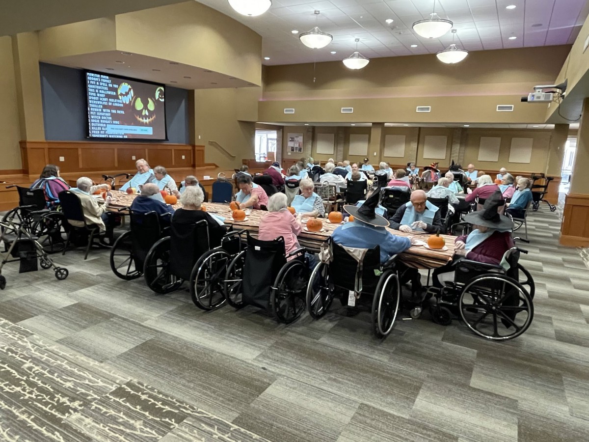 A group of elderly individuals in wheelchairs gathered at tables for a community event in a dining hall.