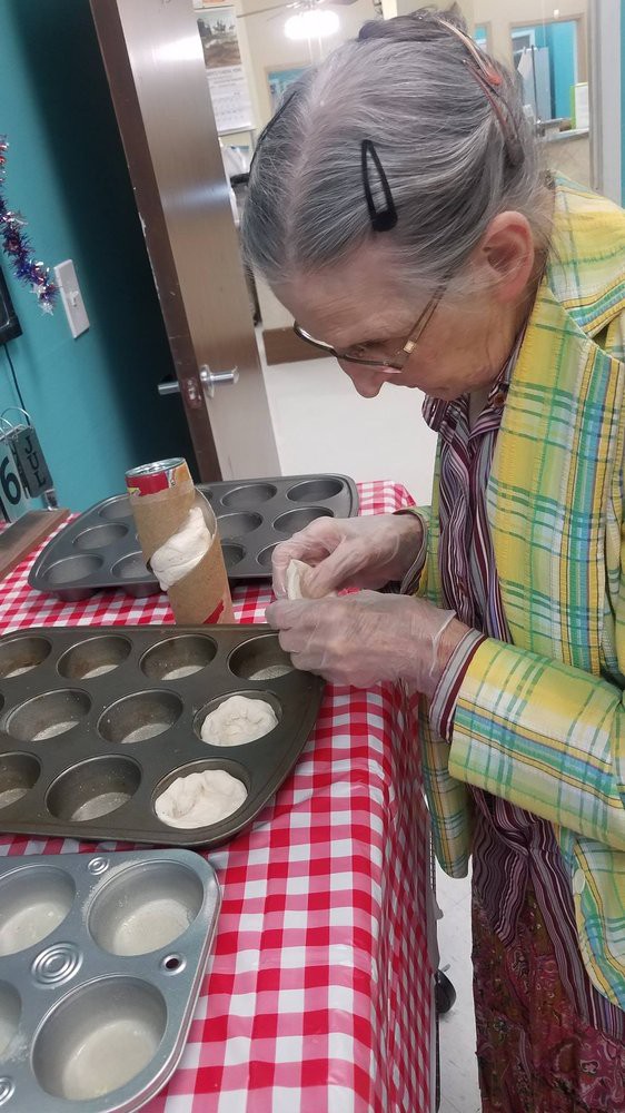 An elderly woman is preparing dough in a muffin tray on a checkered tablecloth.
