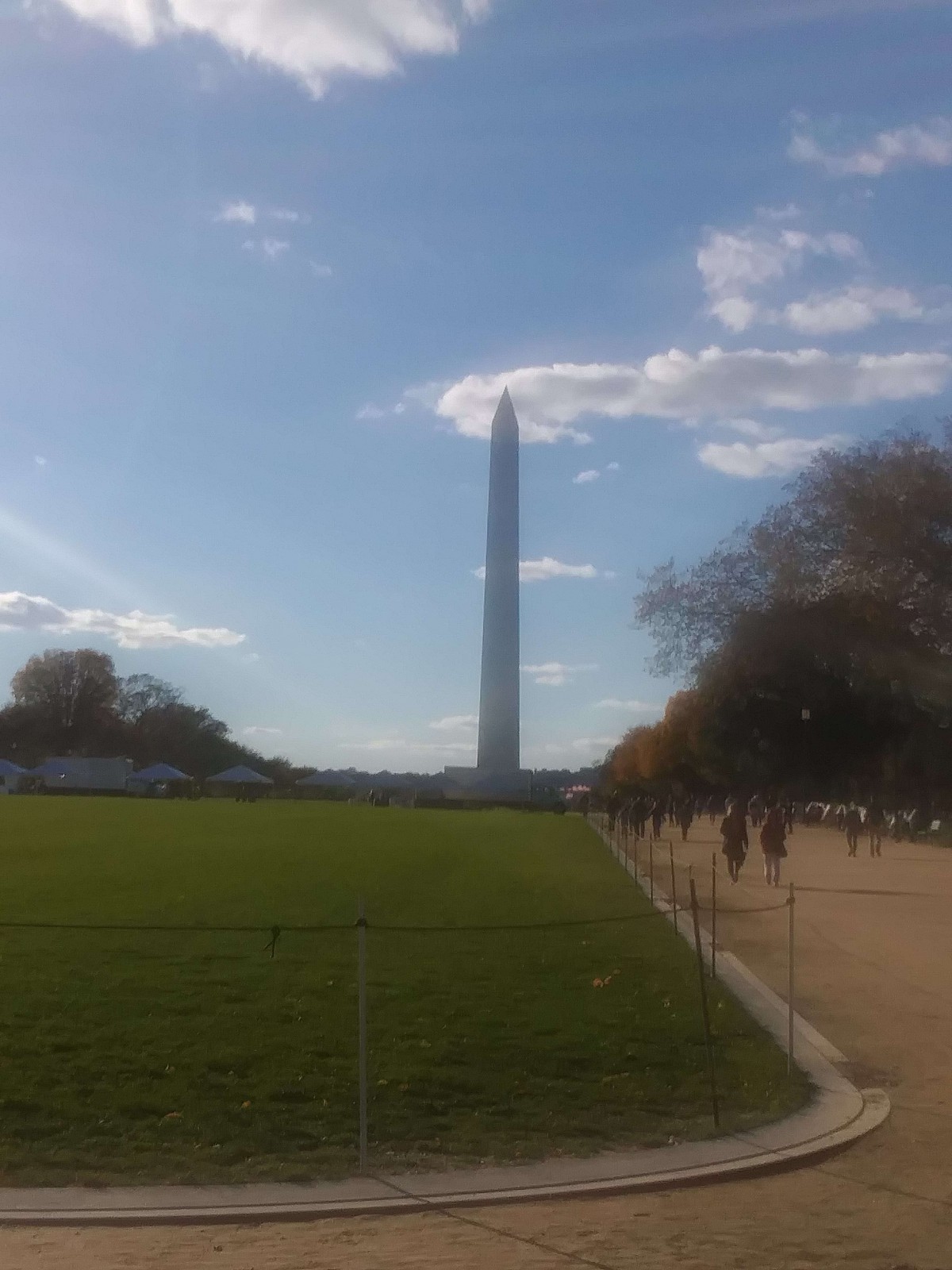 A view of the Washington Monument on a sunny day with clouds in the sky and people walking along a pathway.