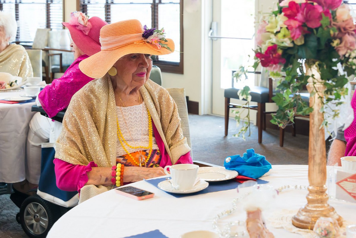 An elegantly dressed elderly woman enjoys tea at a table adorned with a floral arrangement.