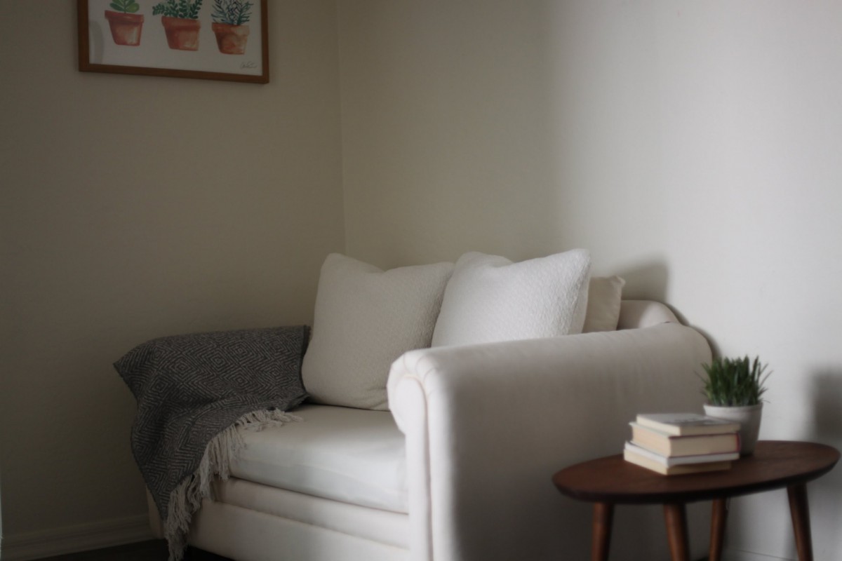 Cozy living room corner with a white sofa, plush pillows, and a side table topped with books.