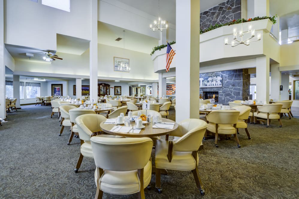 A spacious and elegantly decorated dining area with multiple tables and chairs, featuring chandeliers and a carpeted floor.