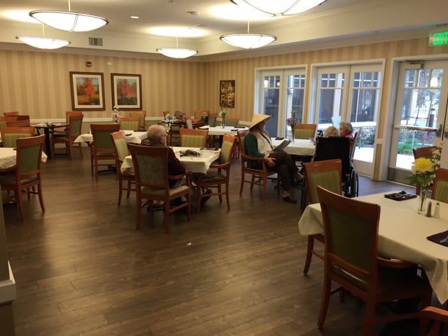 A quiet dining room with tables and chairs, occupied by a few seniors.