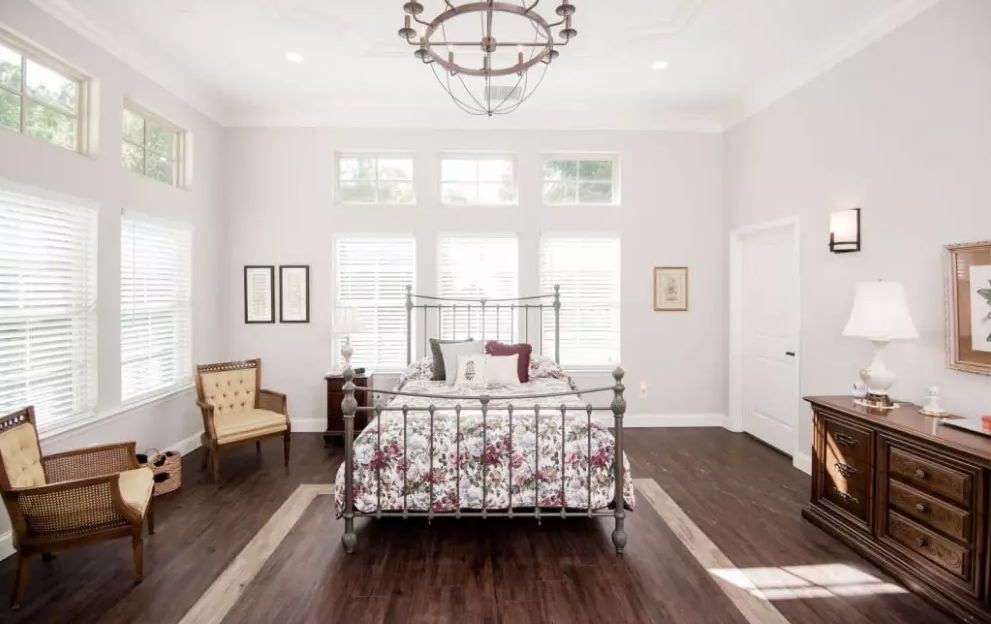 A spacious and elegantly designed bedroom featuring a metal bed frame, floral bedding, and a large chandelier. The room is adorned with wooden flooring and white walls, enhanced by natural light from multiple windows.