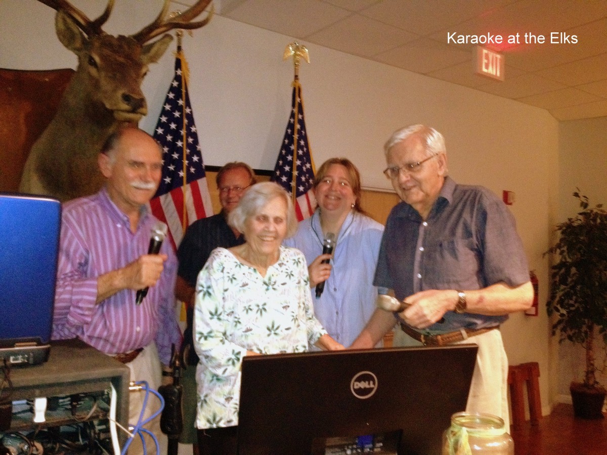 A group of elderly individuals gathered indoors, holding microphones, with American flags in the background.