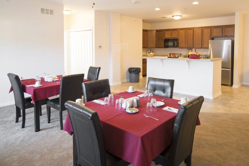 A dining area with tables set for a meal, featuring red tablecloths and leather chairs, adjacent to a kitchen.