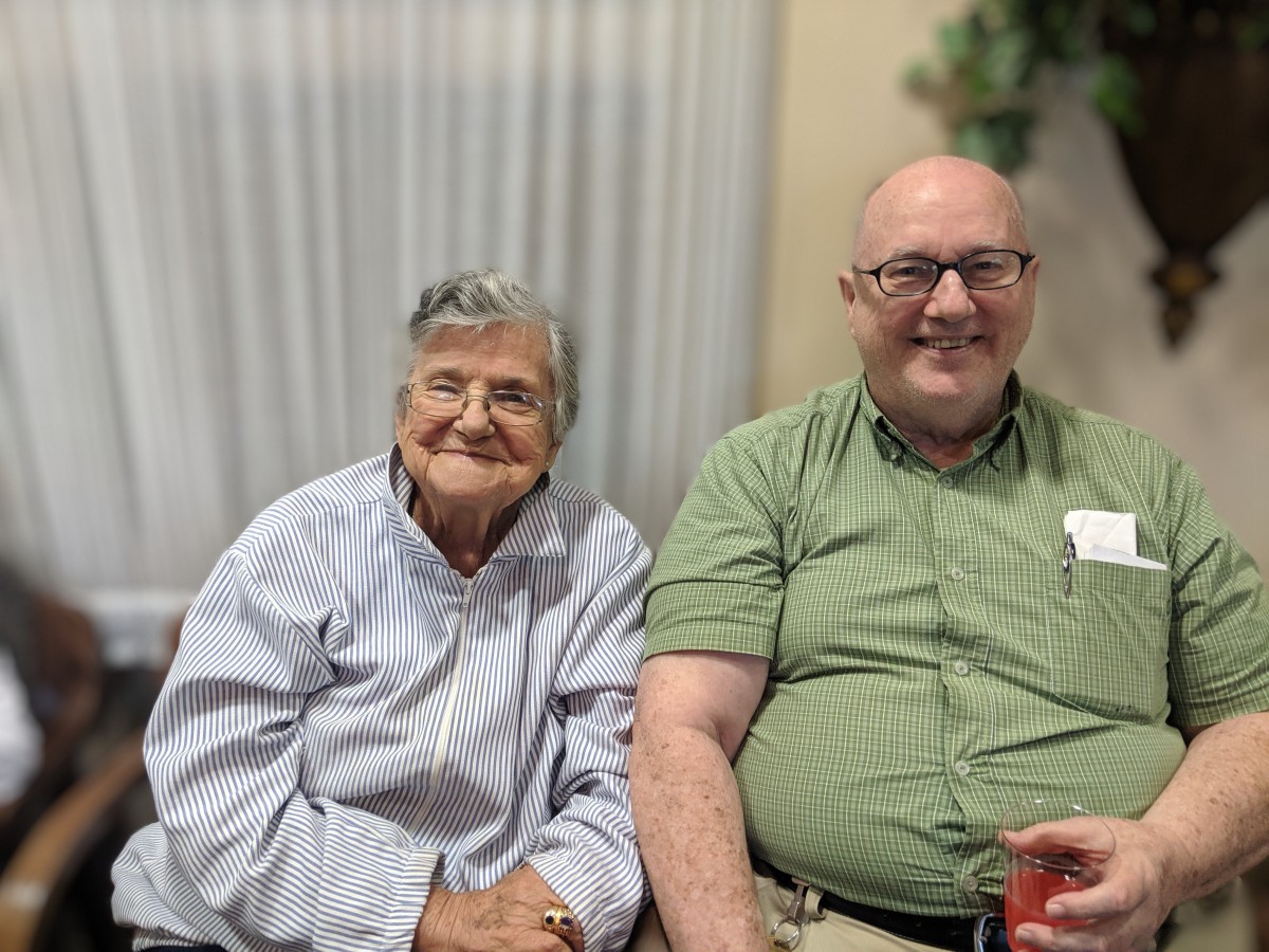 An elderly woman and an adult man sitting together indoors, both smiling.