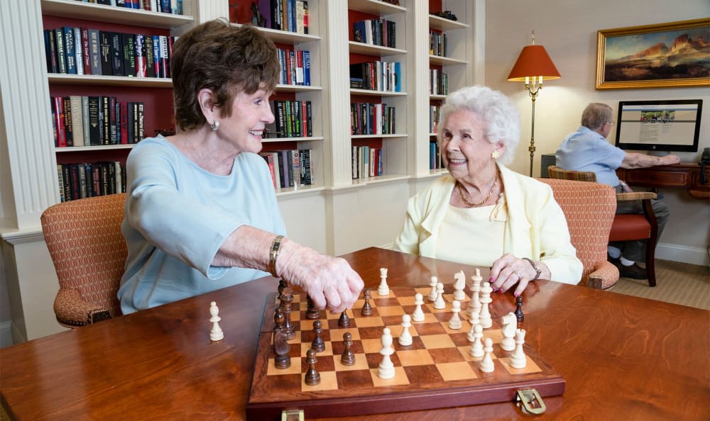 Two elderly women enjoy a game of chess in a cozy library setting.