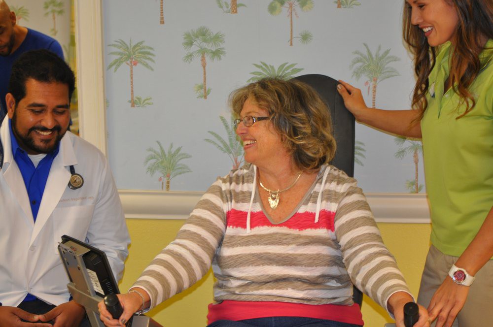 A smiling woman seated indoors, wearing a striped shirt with palm tree wallpaper in the background.