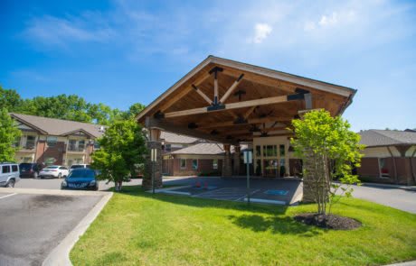 A building entrance with a large wooden canopy, surrounded by trees and a parking lot on a sunny day.