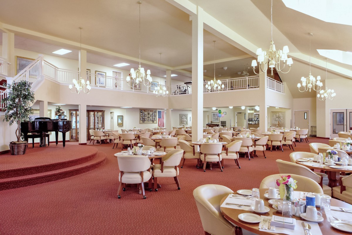 A spacious dining hall with elegantly arranged tables and chairs, featuring chandeliers and a carpeted floor.