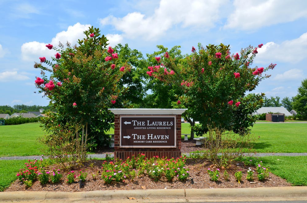 A directional sign surrounded by trees and flowers on a sunny day.