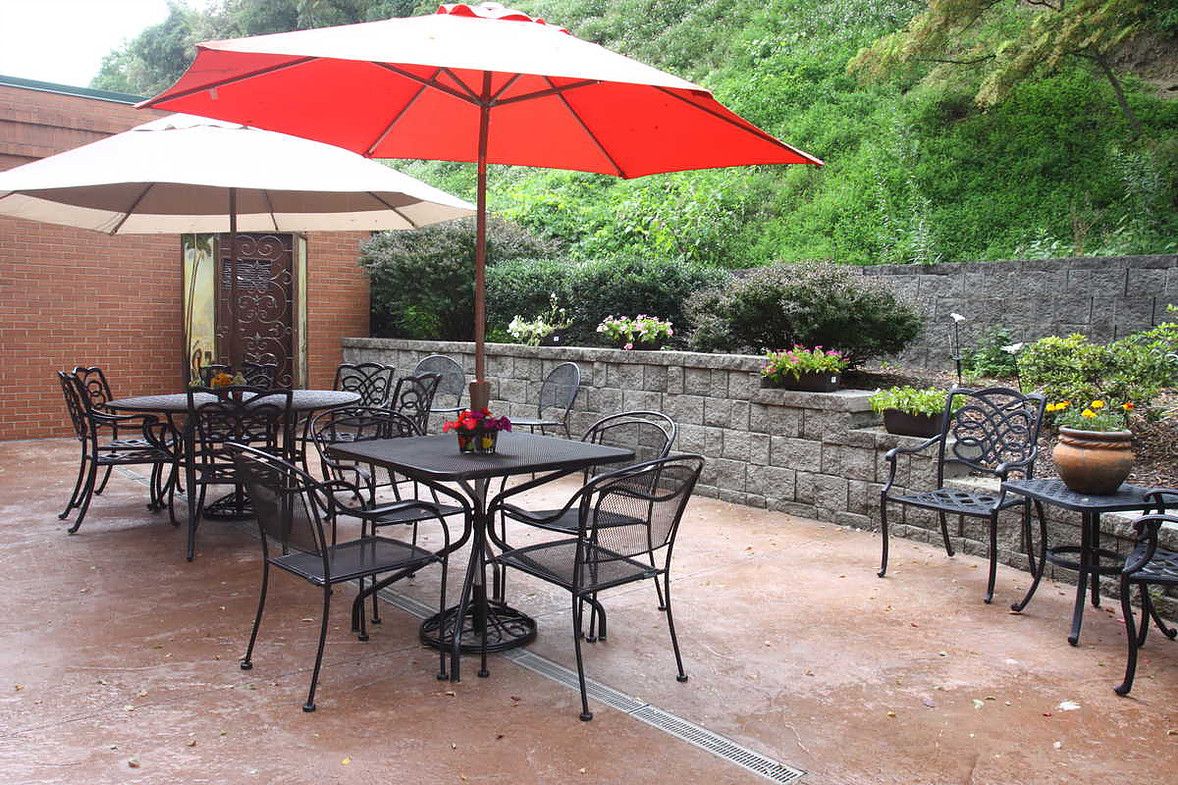 A pleasant outdoor patio with tables, chairs, and red umbrellas surrounded by greenery.