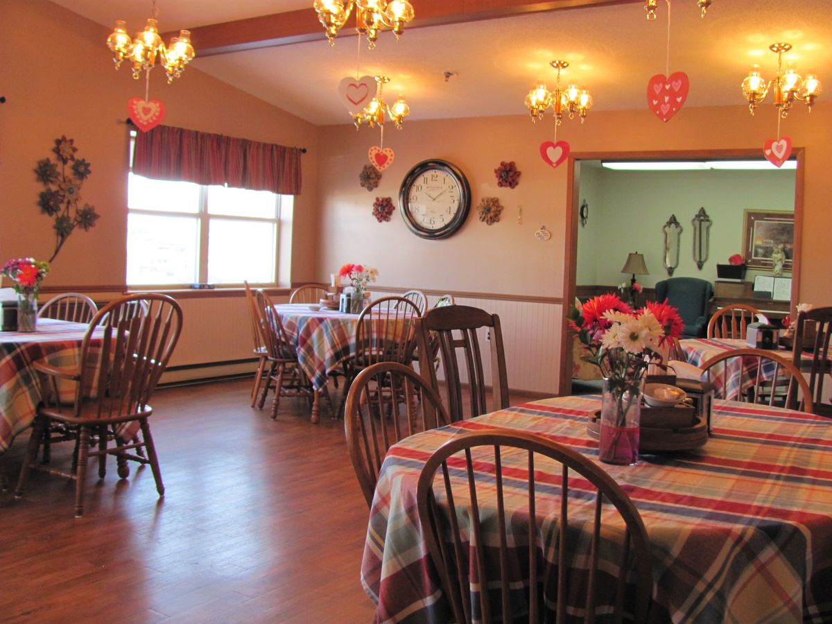 A cozy dining room with decorated tables and wooden chairs, featuring a large clock on the wall.