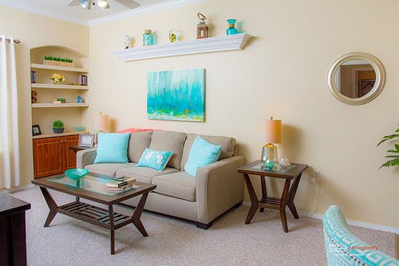 A cozy living room featuring a sofa with teal cushions, a wooden coffee table, decorative shelves, and colorful artwork.