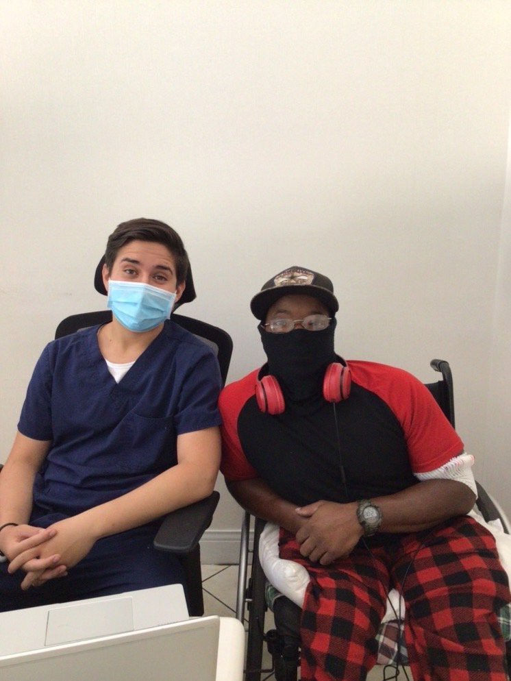 Two people sitting indoors, wearing masks and casual clothing.