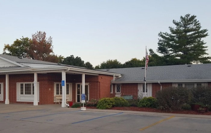 A brick building with a covered entrance and a flagpole, surrounded by trees and parking spaces.