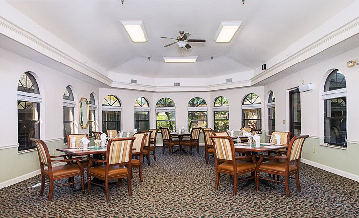 A bright dining room with multiple tables and chairs, featuring large windows and a ceiling fan.