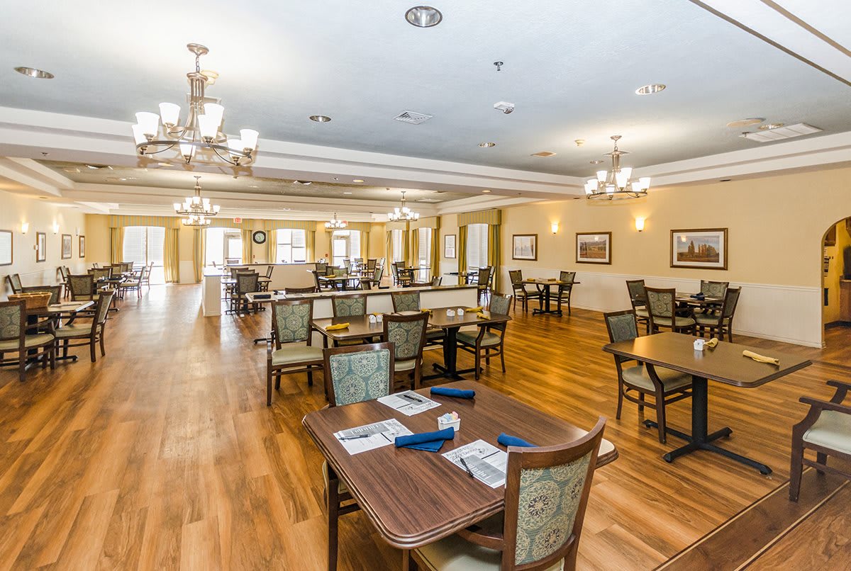A spacious and well-lit dining room with neatly arranged tables and chairs, featuring elegant chandeliers and hardwood flooring.
