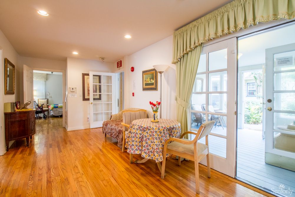 A cozy living room with wooden flooring, featuring a small seating area with a table and chairs by large windows adorned with curtains.