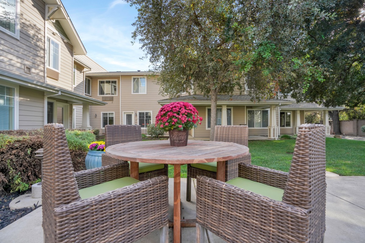 A cozy outdoor patio set with wicker chairs and a table adorned with a pot of vibrant flowers, set in a well-maintained courtyard surrounded by residential buildings.