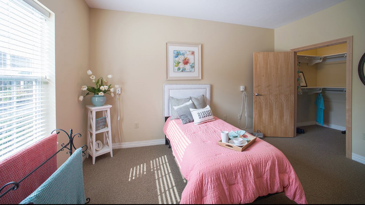 A cozy bedroom with a single bed draped in pink bedding, decorated with pillows and a flower painting on the wall.