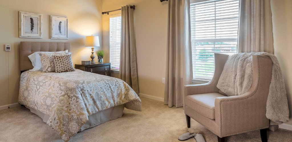 A cozy bedroom with a bed, side table lamp, curtains, and an upholstered chair.