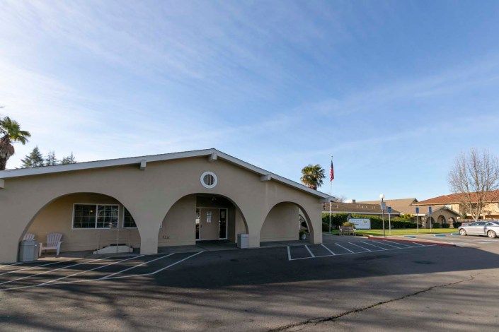 A building with arched entrances and a parking lot in front, featuring a flagpole under a clear sky.