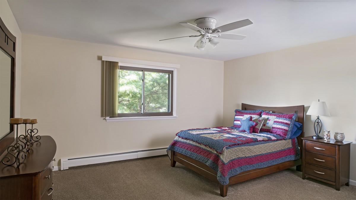 A simple bedroom with a bed, ceiling fan, and a window bringing in natural light.