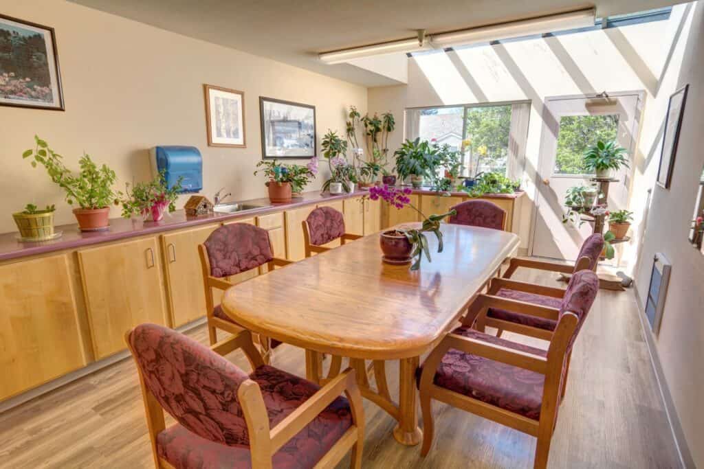 A cozy dining room with a wooden table and chairs, surrounded by indoor plants and sunlight streaming through the windows.