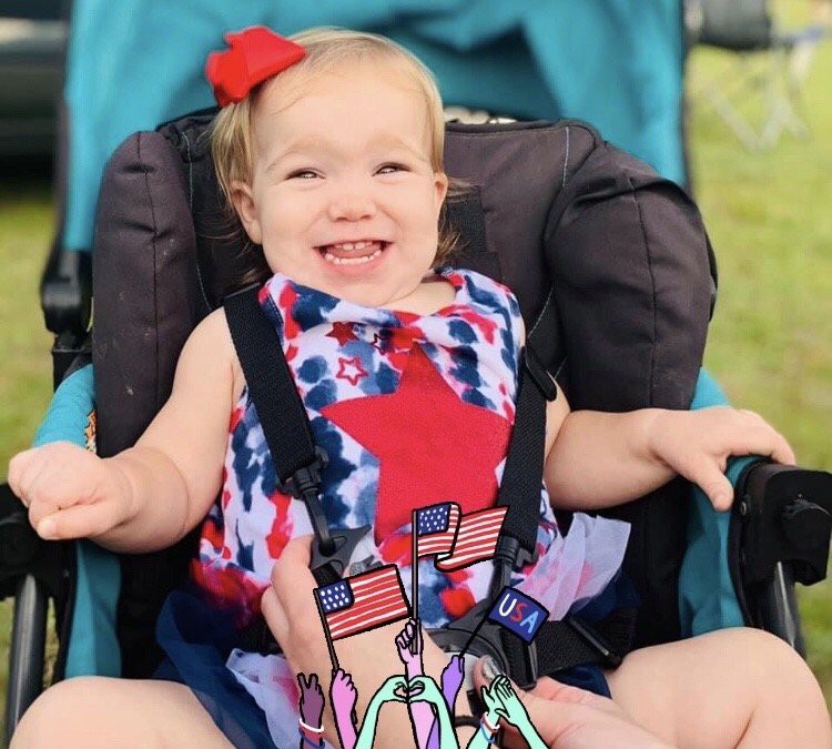 A happy baby wearing a red, white, and blue outfit with a matching bow sits in a stroller.
