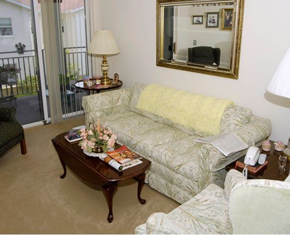A cozy living room featuring a patterned sofa, a wooden coffee table adorned with books and decorations, a large mirror on the wall, and plush carpet flooring.