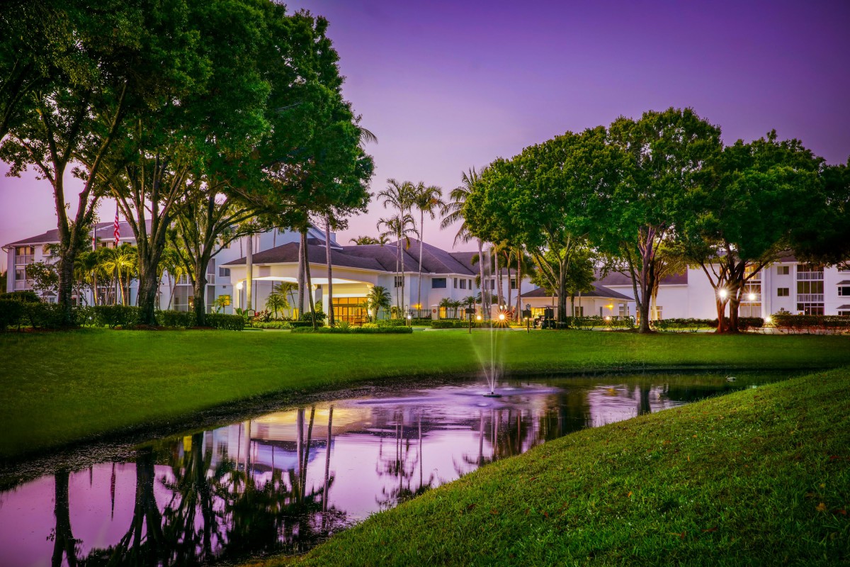 A tranquil evening view of a modern house with its reflection shimmering in a nearby pond, surrounded by lush green grass and trees.