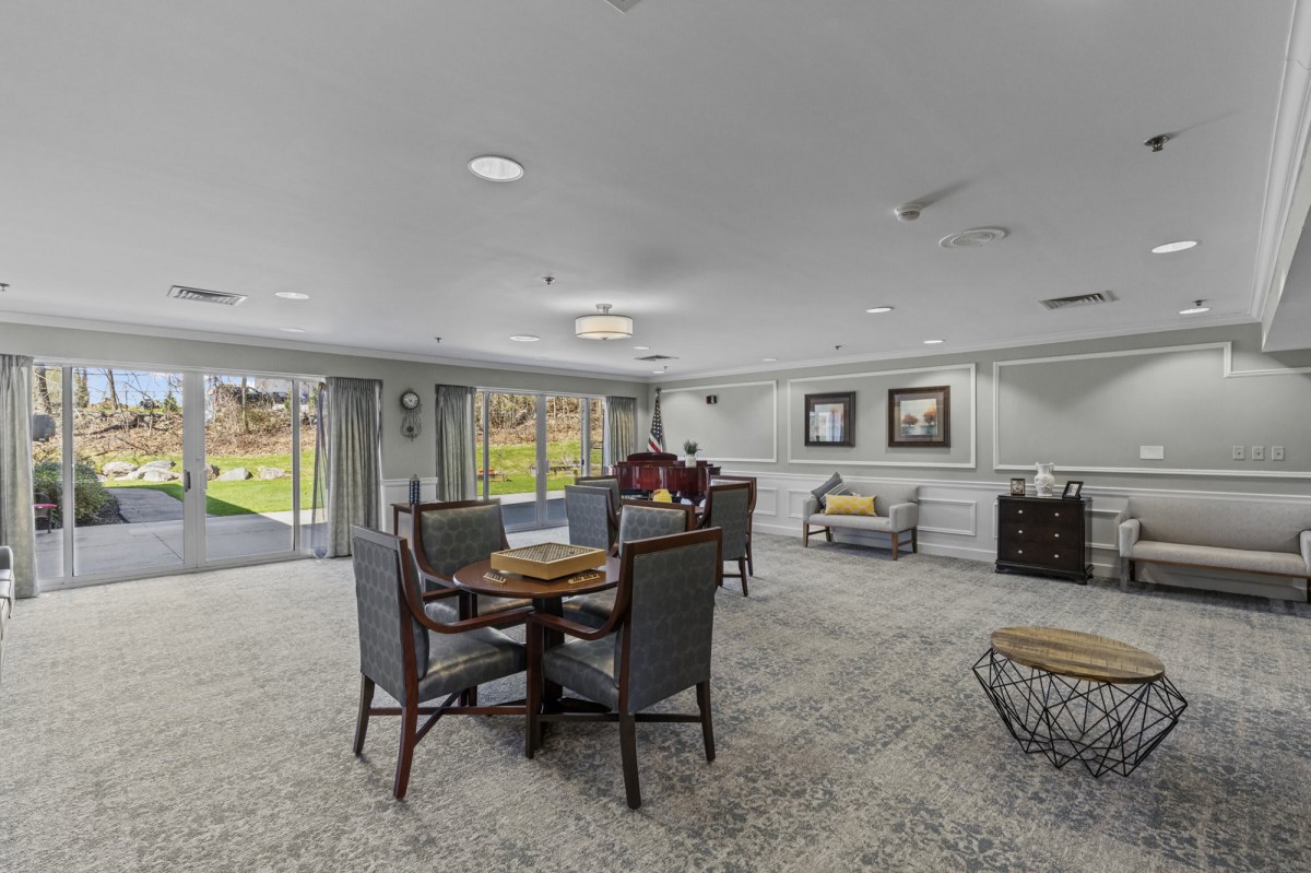 A spacious living room with carpeted flooring, multiple seating areas, and large windows offering a view of the outdoors.