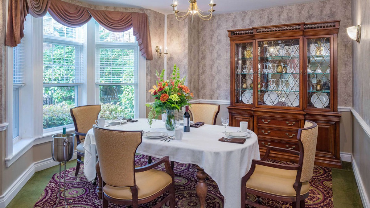 An elegant dining room featuring a set table with a floral centerpiece, surrounded by upholstered chairs and illuminated by a chandelier.
