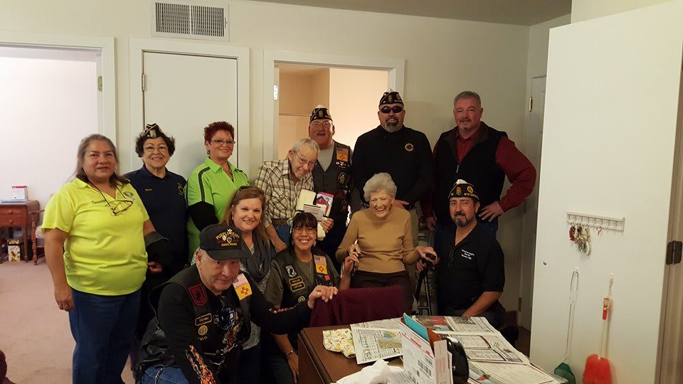A group of people, including veterans wearing military hats, gather around an elderly woman indoors.